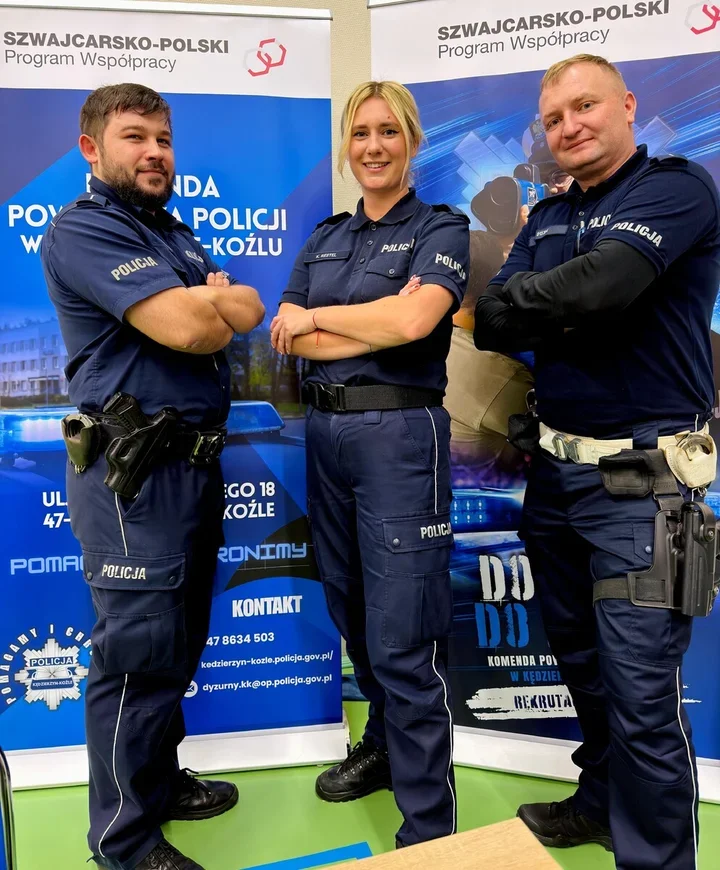 Trzej policjanci w mundurach stoją z założonymi rękami przed banerami Komendy Powiatowej Policji w Kędzierzynie-Koźlu i Szwajcarsko-Polskiego Programu Współprac