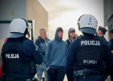 Funkcjonariusze polskiej policji w kaskach i kamizelkach z napisem POLICJA stoją przed grupą mężczyzn.